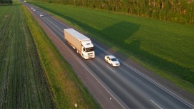 A white truck drives on a country road, green fields can be seen to the right and left of the road. Other vehicles can be seen in front of and behind the truck.
