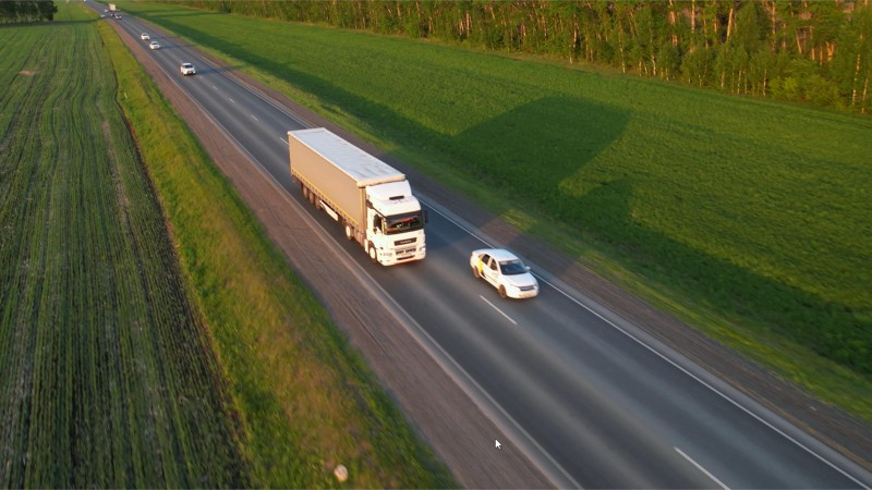 A white truck drives on a country road, green fields can be seen to the right and left of the road. Other vehicles can be seen in front of and behind the truck.