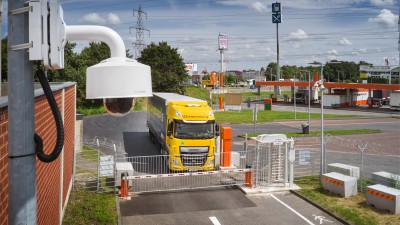 A yellow truck stands in front of a barrier to a security parking lot from Bosch Secure Truck Parking at the Frechen truck stop. A camera can be seen in the foreground.